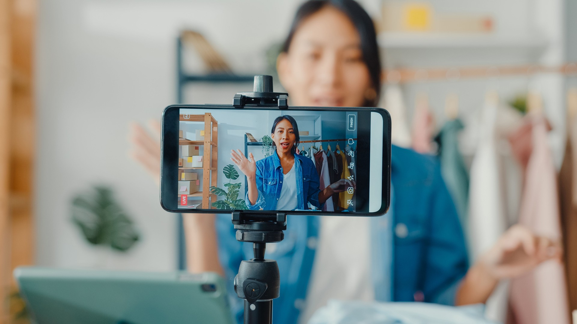 Joven diseñadora de moda de Asia usando el teléfono móvil recibiendo órdenes de compra y mostrando ropa grabando video en vivo transmitiendo en línea en la tienda.