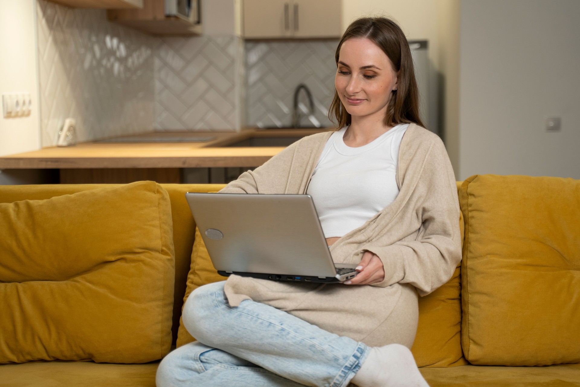 Una mujer joven con ropa informal está sentada en un sofá amarillo usando una computadora portátil, divirtiéndose en las redes sociales, relajándose, sonriendo y riendo. Una mujer freelancer dedicada al marketing de ventas en línea.